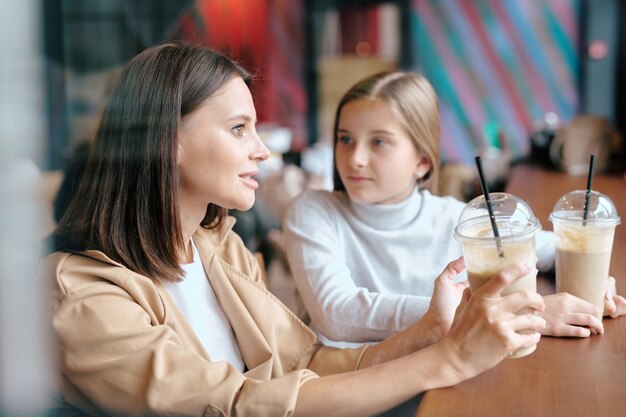 カフェでテーブルに座って飲み物を飲んでいる間彼女のかわいい娘に話しているミルクカクテルを持つかなり若い女性