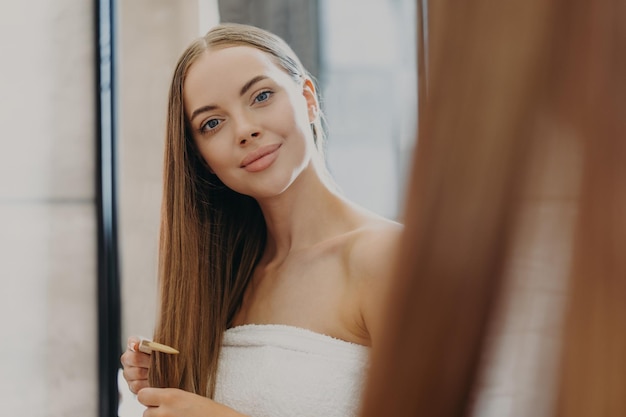 Photo pretty young woman with minimal makeup full lips looks at herself in mirror combs long straight hair wrapped in bath towel cares about herself wants to have fabulous look hair care concept