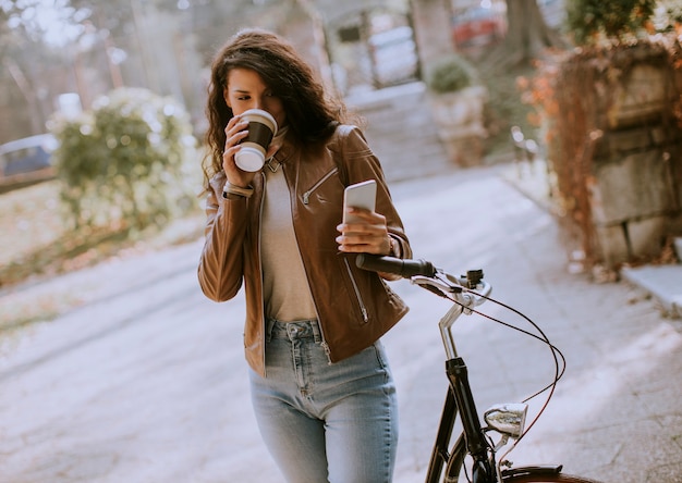 Photo pretty young woman with mobile phone drink coffee to go by the bicycle on autumn day