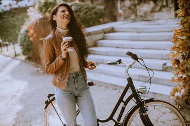 Photo pretty young woman with mobile phone drink coffee to go by the bicycle on autumn day