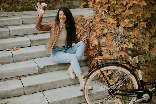 写真 携帯電話を持ったかなり若い女性が秋の日に自転車で階段に行くためにコーヒーを飲む