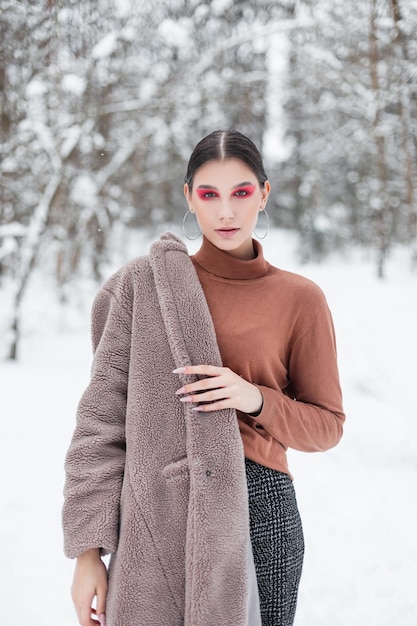 Pretty young woman with pink color makeup in fashion winter\
clothes with vintage sweater and coat in a snowy park