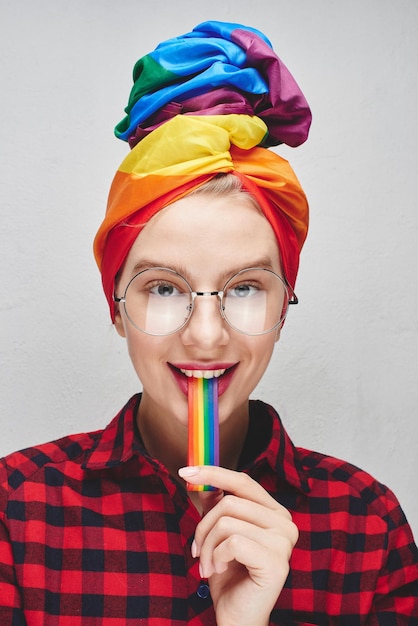 Una bella giovane donna con occhiali rotondi e labbra carnose in un turbante e una maglietta rossa tiene un arcobaleno in bocca contro un muro bianco. lgbtq lesbiche, gay, bisessuali, transgender, queer. uomo omosessuale