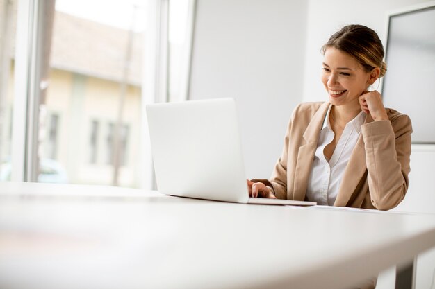 Pretty young woman working on laptop in bright office with big screen behind her