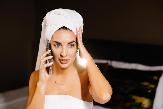 Pretty young woman wrapped in a shower towel using mobile phone while sitting on bed and talking to phone