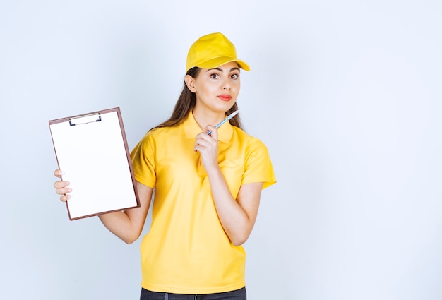 Pretty young woman in yellow courier uniform with clipboard posing over white wall.