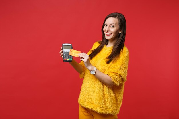 Pretty young woman in yellow fur sweater hold wireless modern bank payment terminal to process and acquire credit card payments isolated on red background. People sincere emotions, lifestyle concept.