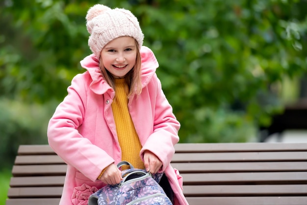 Prety school girl kid with backpack sitting on the banch at autumn park and smiling beautiful female