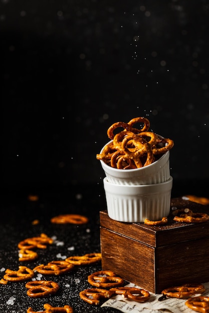 Pretzels lie in two cups on a dark background. A large amount of salt is poured on top of the bretzel and scattered around.