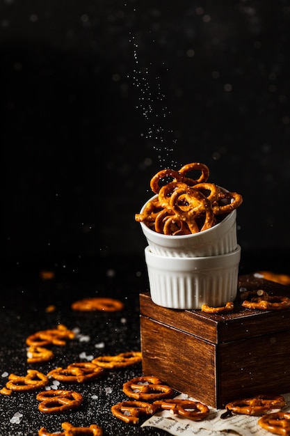 Pretzels lie in two cups on a dark background. A large amount of salt is poured on top of the bretzel and scattered around.