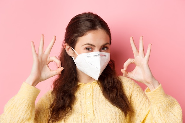 Preventive measures, health care concept. Young glamour girl wearing medical respirator during covid-19 pandemic, showing okay signs, praise excellent choice, recommends something.