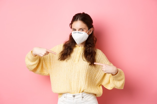 Preventive measures health care concept young happy woman protect herself from covid with medical respirator points at her chest shows your logo or promotional text pink wall