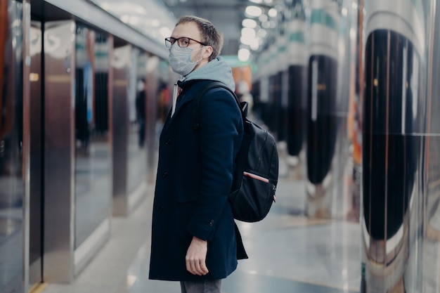 Preventive measures in public places Sideways shot of man in medical mask stands on metro station carries backpack looks worried during epidemic situation People prevention coronavirus