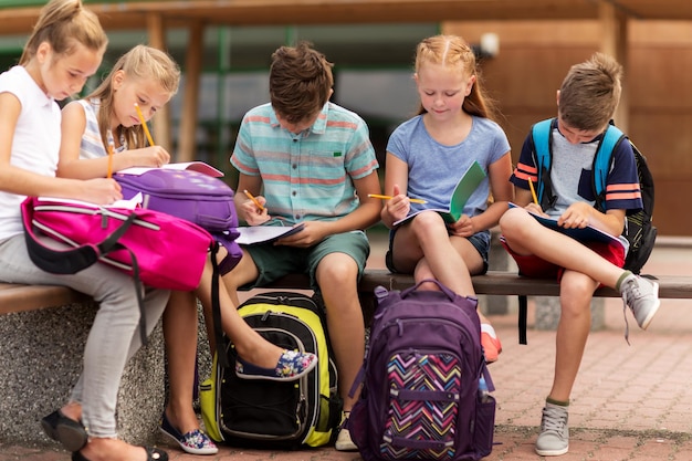 primary education, friendship, childhood, communication and people concept - group of happy elementary school students with backpacks and notebooks sitting on bench and doing homework outdoors