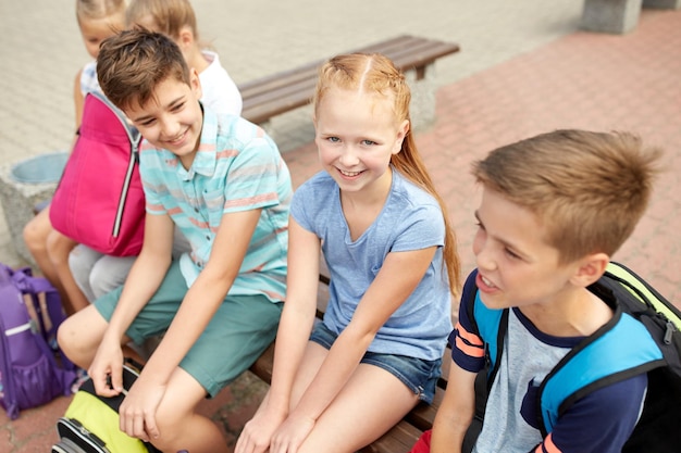 primary education, friendship, childhood, communication and people concept - group of happy elementary school students with backpacks sitting on bench and talking outdoors