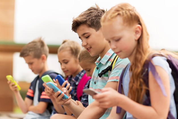 начальное образование, дружба, детство, технологии и концепция людей - группа счастливых учеников начальной школы со смартфонами и рюкзаками, сидящих на скамейке на открытом воздухе