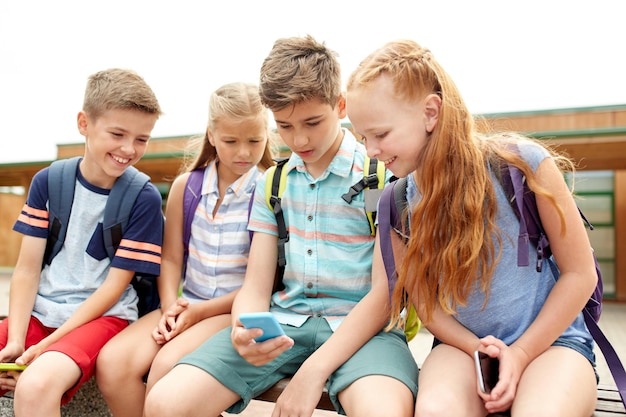 primary education, friendship, childhood, technology and people concept - group of happy elementary school students with smartphones and backpacks sitting on bench outdoors