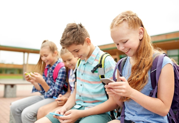 primary education, friendship, childhood, technology and people concept - group of happy elementary school students with smartphones and backpacks sitting on bench outdoors