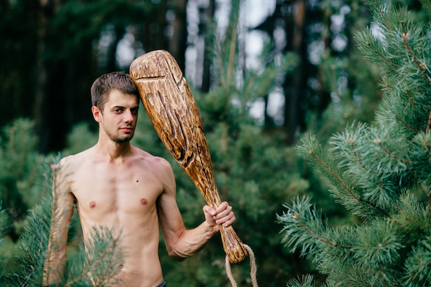 Primitive naked man with huge wooden stick posing in forest