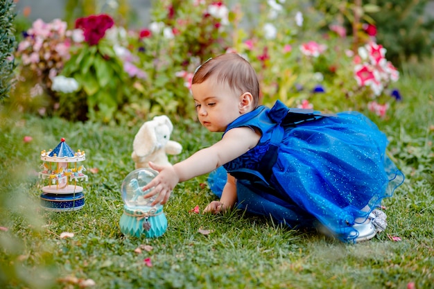 Princess baby girl with blue dress sitting on grass in garden and playing with one of her toys