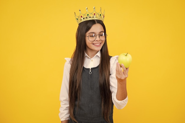 Princess child in queen crown hold apple isolated on yellow background Teenage girl wear diadem Happy girl face positive and smiling emotions