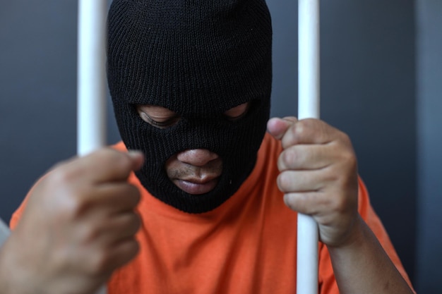 a prisoner in orange shirt and black mask inside the bars of a prison