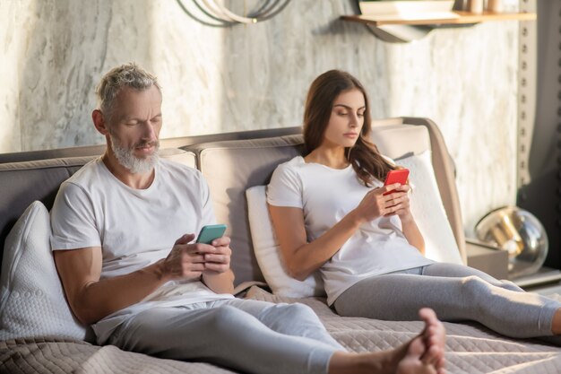 Privacy. Serious busy man and woman in home clothes with smartphones sitting at home on bed