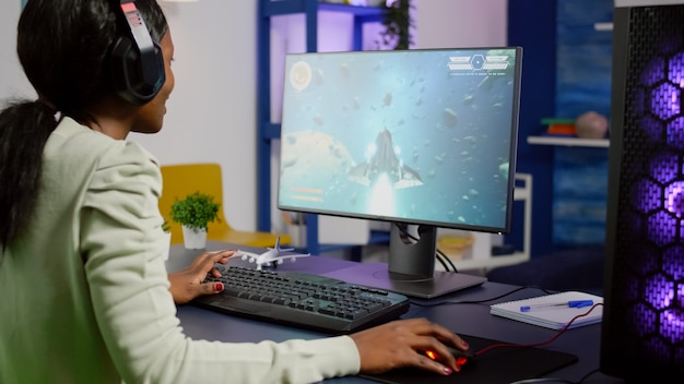 Pro black woman gamer playing space shooter videogame on RGB powerful computer wearing professional headphones