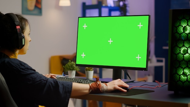 Pro woman player gaming on powerful computer with green mock up chroma key screen while streaming online competition. Gamer using pc with greenscreen isolated desktop streaming shooter games