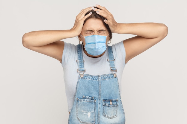 Probleem, crisis, slecht nieuws of hoofdpijn. Portret van een geërgerde jonge brunette vrouw met een chirurgisch medisch masker in een denim overall die haar hoofd vasthoudt. indoor studio-opname geïsoleerd op een grijze achtergrond