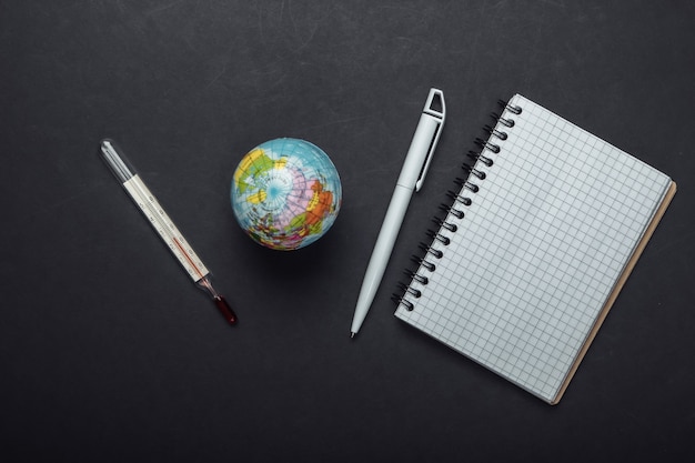 The problem of global warming. Globe, thermometer with a notebook on black background. Top view