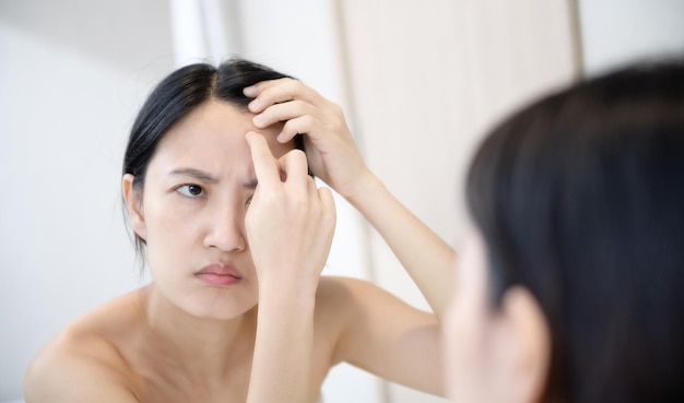 Problem skin Concerned young asian women popping pimple on cheek while standing near mirror in bathroom young asian women with acne