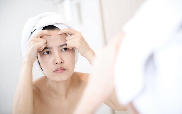 Problem skin Concerned young asian women popping pimple on cheek while standing near mirror in bathroom young asian women with acne