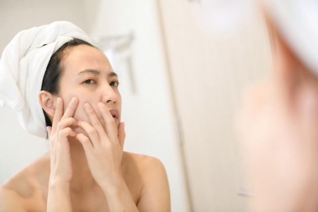 문제 피부 여드름을 가진 젊은 아시아 여성 화장실에서 거울 근처에 서있는 동안 뺨에 여드름을 터뜨리는 문제 피부