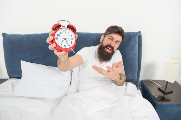 Problem with early morning awakening Get up with alarm clock Overslept again Tips for waking up early Tips for becoming an early riser Man bearded hipster sleepy face in bed with alarm clock