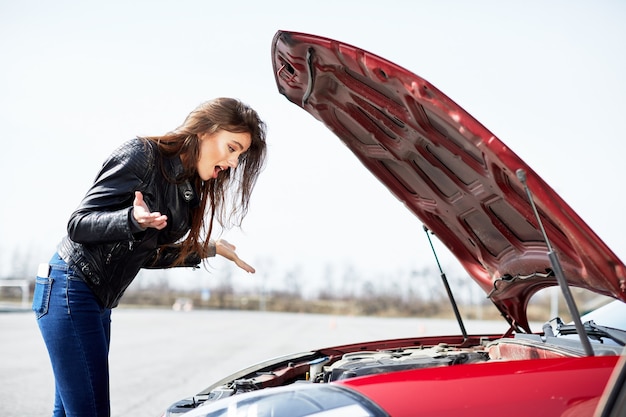 Foto problemi con il motore, conducente confuso. profilo di donna stressata in piedi vicino all'automobile. guardando dentro la macchina rossa rotta, fuori