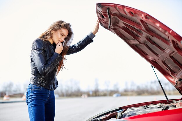 エンジンの問題、混乱したドライバー。車の近くに立っている女性のプロフィール、分解。赤い車の中、外を見る