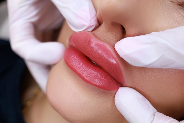 Процедура перманентного макияжа губ с помощью машинки мастер нежно проводит пальцами по губам модели