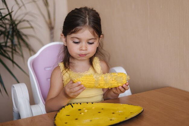 Proces van een klein meisje dat verse cornat eet, een schattig kind zit op tafel en eet maïs