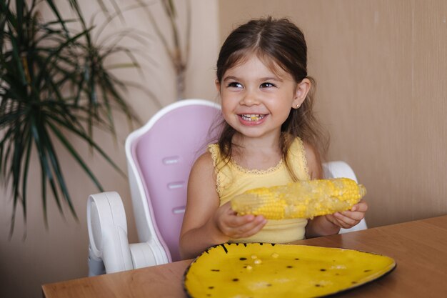 Proces van een klein meisje dat verse cornat eet, een schattig kind zit op tafel en eet maïs