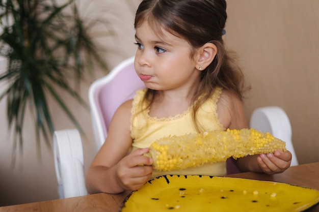 Proces van een klein meisje dat verse cornat eet, een schattig kind zit op tafel en eet maïs