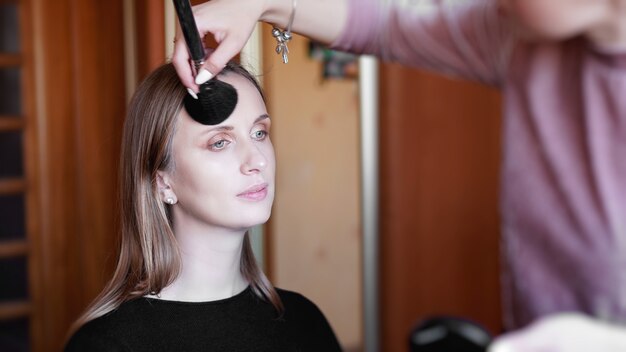 Proces van het maken van make-up. Visagist werken met penseel op model gezicht. Portret van jonge vrouw in interieur.