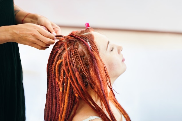 Proces van vlechten vlechten op hoofd in schoonheidssalon close-up roodharig meisje maakt vlechten dreadlocks dr...