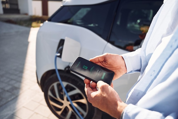 Foto il processo di ricarica viene visualizzato sullo smartphone vista ravvicinata dell'uomo con la sua auto elettrica