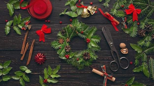 Process of creating Christmas wreath with various tools and trinkets on wooden background, top view