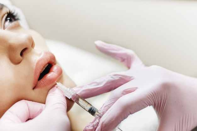 The process of lips enhancement. Cosmetician makes an injection of hyaluronic acid in a beautiful lips. The young girl with a beautiful face in the special hat and hands of the doctor in the pink glov