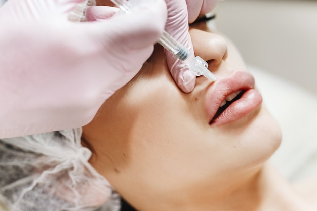 The process of lips enhancement. Cosmetician makes an injection of hyaluronic acid in a beautiful lips. The young girl with a beautiful face in the special hat and hands of the doctor in the pink glov