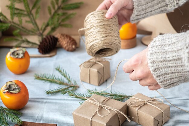 The process of packaging stylish modern gifts for Christmas and New year. Gift boxes made of Kraft paper, twine and Christmas tree branches. Christmas background, holiday atmosphere.