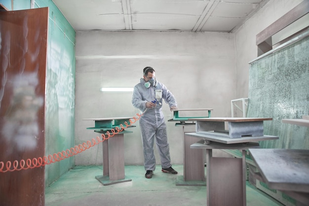 Process of production and manufacture of wooden furniture in\
factory worker man in overalls paints wood with paint spray