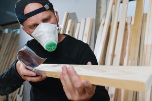 Foto lavorazione del legno per la fabbricazione di mobili un falegname in abiti scuri che indossa un respiratore macina le tavole con carta vetrata